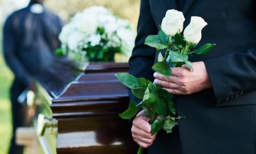 Close-up of matue man in black suit holding two fresh white roses while standing in front of camera against coffin with closed lid